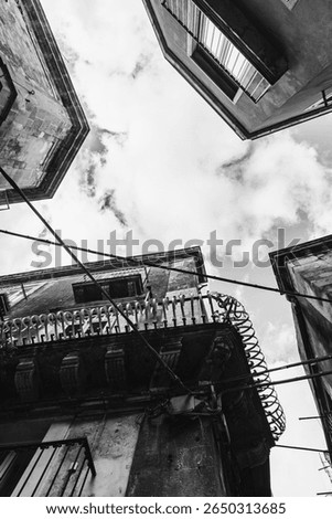 A dramatic black and white upward view of old Sicilian buildings with balconies, wires, and cloudy sky.