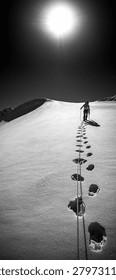 A dramatic black and white vertical shot of a lone mountaineer ascending a steep, sun-drenched snow slope in the Wildspitze region of Tyrol, Austria, with a climbing rope stretching along deep footprints towards a burning sun.