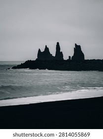 Dramatic black and white view of Víkurfjara Beach in Vík, Iceland, with the iconic Reynisdrangar sea stacks, also known as the Troll Fingers, rising from the Atlantic Ocean in the background. Dark volcanic sand, powerful waves and rainy, stormy weather create a moody and atmospheric Icelandic coastal landscape.