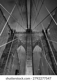 Dramatic black and white view looking upward at the iconic suspension cables and stone towers of the Brooklyn Bridge, showcasing geometric symmetry and architectural detail against a moody sky.