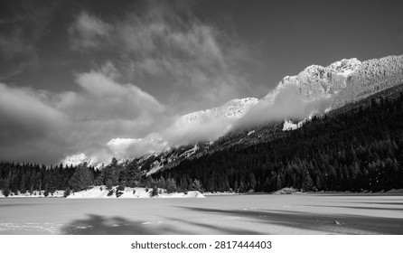 Dramatic black and white winter landscape of snow covered mountain peaks wrapped in clouds above a frozen lake and evergreen forest in the North Cascades of Washington