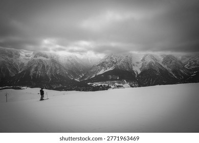 Dramatic black and white winter mountain landscape featuring a lone skier on a vast snowy slope beneath dark storm clouds. The panoramic alpine scenery showcases rugged snow-covered peaks, deep valleys, and a moody monochrome atmosphere, evoking solitude, adventure, and the raw beauty of nature. Perfect for themes such as winter sports, alpine travel, wilderness exploration, and minimalist outdoor photography.