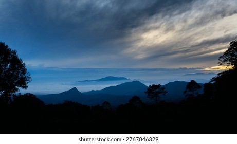 Dramatic blue hour mountain landscape with layered hills and misty valleys under a cloudy sky of West Java area, Indonesia. Silhouetted trees frame the foreground, creating a serene and atmospheric scene that captures the natural beauty and tranquility of Indonesia’s highland regions at dawn.