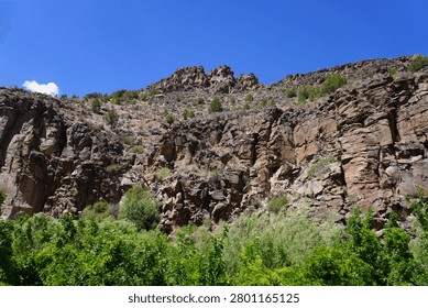 A dramatic canyon view looking up at massive, rugged rock cliffs and steep mountain slopes dotted with green trees under a vibrant blue sky.