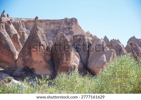 Dramatic Cappadocia landscape featuring towering fairy chimneys, eroded tuff pillars with ancient cave dwellings carved into cliffs. Arid terrain dotted with green bushes under endless blue sky. Ideal for tourism ads, adventure travel campaigns, UNESCO heritage visuals, hiking posters, geological wonders books. Captures timeless Anatolian magic boosting wanderlust sales.
