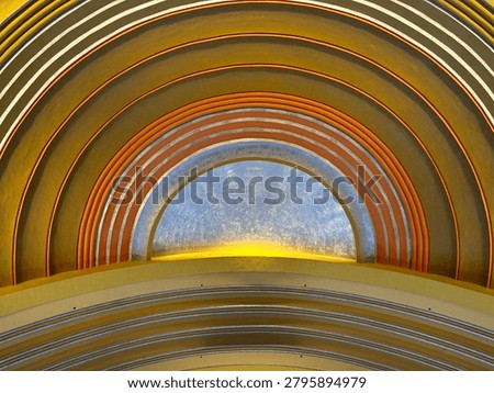 Dramatic ceiling fresco-type artwork and sky painting, with gilded edges and sweeping architecture, in a train station from its glory days, share artistic care and meticulous work high above the floor capturing grandeur of the human spirit and artistic pursuits. Great for backgrounds, room for text, textures, backdrops.- A large half-circle shape dominates the image.
Multiple concentric orange and white bands or ribs arch over the top.
The center section is a bluish-gray textured surface, possibly metal, frosted glass, or painted material.
A warm yellow light glows along the bottom edge of the semicircle, creating a backlit effect.
The overall appearance resembles:
An illuminated archway or alcove,
The inside of a decorative dome,
A retro-futuristic ceiling feature,
Or a stylized architectural element in a building or transportation hub.
The image is cropped tightly enough that it's difficult to determine the exact object or location, but it gives a strong impression of curved architectural design with integrated lighting and layered arches.
