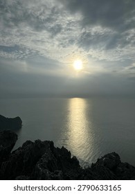 Dramatic cinematic sunset over the ocean with sun rays reflecting on the water surface and dark rocky foreground.⁠