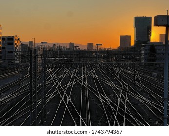 Dramatic cityscape featuring a complex railway network at sunset, with multiple train tracks converging beneath a glowing orange sky. A commuter train travels through the urban rail yard, surrounded by overhead power lines and modern high-rise buildings silhouetted against the setting sun. The scene captures transportation infrastructure, urban mobility, and industrial architecture during golden hour.