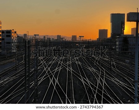 Dramatic cityscape featuring a complex railway network at sunset, with multiple train tracks converging beneath a glowing orange sky. A commuter train travels through the urban rail yard, surrounded by overhead power lines and modern high-rise buildings silhouetted against the setting sun. The scene captures transportation infrastructure, urban mobility, and industrial architecture during golden hour.
