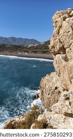 A dramatic cliff face frames a view of the Mediterranean Sea, a pebble beach with colorful cabanas, and rolling hills dotted with white buildings under a clear sky