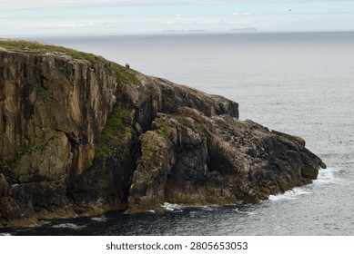 Dramatic cliff face on the Isle of Lewis, Outer Hebrides, Scotland, with distant islands visible on the horizon across the Atlantic Ocean. The vertical Lewisian gneiss cliff shows characteristic columnar jointing and lichen colouring. A Blackface sheep is visible on the clifftop edge, giving scale to the dramatic drop.