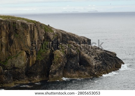 Dramatic cliff face on the Isle of Lewis, Outer Hebrides, Scotland, with distant islands visible on the horizon across the Atlantic Ocean. The vertical Lewisian gneiss cliff shows characteristic columnar jointing and lichen colouring. A Blackface sheep is visible on the clifftop edge, giving scale to the dramatic drop.