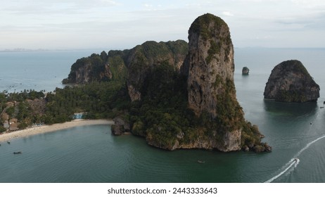 Dramatic cliffs draped in tropical foliage rise above a serene beach with a bustling coastal village, as boats trace white lines through the jade-green sea waters. - Powered by Shutterstock