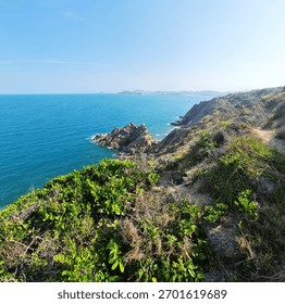 Dramatic cliffside view with a hiking path, overlooking the vast blue ocean and a cluster of rugged, jagged rock formations offshore