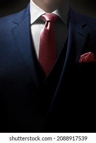 A dramatic close up shot of a man dressed in an expensive formal suit. The jacket is navy blue with a matching waistcoat as well as a pink pocket square and tie.