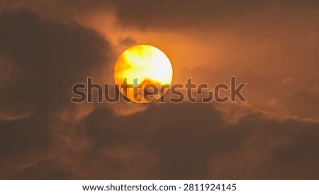 Dramatic close-up of a large glowing golden sun or full moon partially hidden behind dark, moody clouds during a misty sunset or sunrise