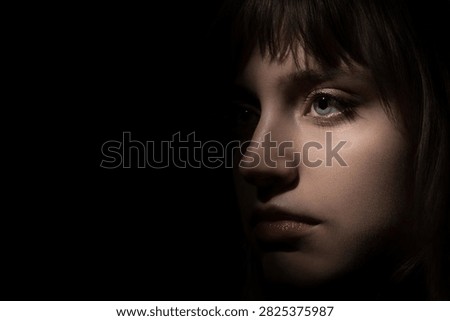 A dramatic close-up low-key portrait of a young woman with striking blue eyes emerging from complete darkness. The narrow light beam highlights one side of her face creating high contrast.