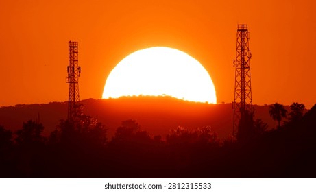 Dramatic close-up of a massive orange sun setting behind hills with silhouettes of radio antennas and forest trees, Cyprus