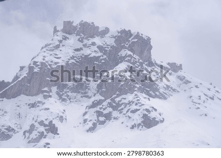 A dramatic close-up of a massive, rugged rocky mountain peak covered in winter snow during a heavy whiteout blizzard.