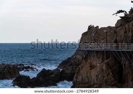 A dramatic close-up morning view of a structured wooden pedestrian deck securely attached to a towering, vertical rock cliff with wild ocean waves crashing far below in Samcheok.