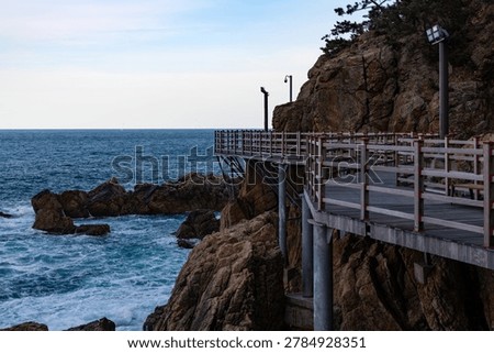 A dramatic close-up morning view of a structured wooden pedestrian deck securely attached to a towering, vertical rock cliff with wild ocean waves crashing far below in Samcheok.