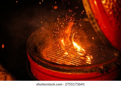 A dramatic close-up of a red ceramic grill with flames and sparks bursting from the grates. The lid is partially open, revealing the intense heat and smoky ambiance — perfect for barbecue-themed stock imagery.