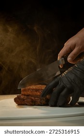 A dramatic, close-up shot of a chef's hands—one bare and one in a black nitrile glove—slicing through a steaming, golden-brown roasted pork belly with a sharp kitchen knife. Thick, warm steam rises from the meat against a dark background, highlighting the contrast between the crunchy crackling skin and the tender layers of meat. The scene captures the final preparation of a perfectly cooked, savory dish.