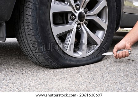 A dramatic close-up of an unrecognizable man's hand holding a sharp knife near a flat car tire, illustrating a concept of road rage, parking conflict, or targeted vandalism. This powerful image captures the moment of a deliberate tire puncture, representing criminal activity, revenge, or a dangerous street encounter. Ideal for articles on automotive security, insurance claims related to intentional damage, crime news, and legal services. The visual of a cold steel blade against the rubber wheel highlights themes of aggression, sabotage, and the breakdown of safety in urban environments. Whether used for a thriller-themed project or a real-world safety guide, this shot effectively conveys a sense of threat, property damage, and anonymous antisocial behavior in a parking lot or on the roadside.