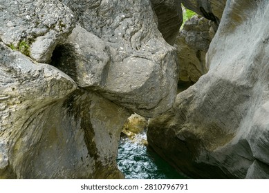 Dramatic close-up view through massive, weathered gray and brown boulders in a natural rocky gorge. The rugged rock formations frame a striking glimpse of vibrant emerald-green water flowing below, with sunlight illuminating the textured stone surfaces and small patches of vegetation growing on the rocks.