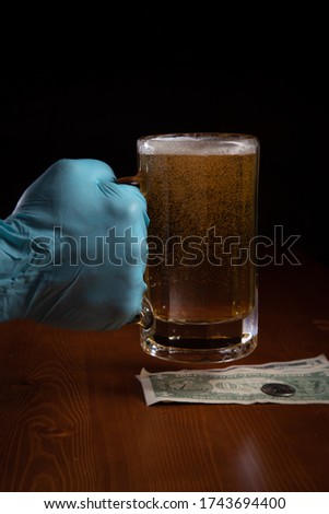 Dramatic close-ups of gloved hands of a bartender or patron at a bar or restaurant, during quarantine, handling a glass/mug of beer with money against a wooden bar top with a black background. 