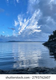 Dramatic Cloud Formation Over Calm Coastal Waters with Reflections in Norway. Serene coastal landscape in this small town Bjørnafjorden, featuring dramatic cloud formations reflected in calm blue water. Rocky shoreline, pine trees, and distant islands create a peaceful Scandinavian seascape with ample copy space for travel, nature, and environmental themes.