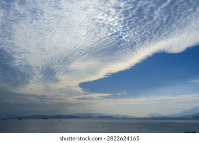 Dramatic cloud formation over a calm sea with distant mountain silhouettes and small boats, creating a tranquil coastal landscape under a blue sky.