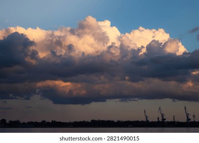 Dramatic cloud formations above the Daugava River in Riga, Latvia, featuring silhouettes of cranes and a distant waterfront landscape during sunset
