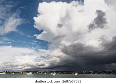 Dramatic cloud formations over a coastal landscape with boats and a bridge, showcasing the beauty of nature's contrasts.