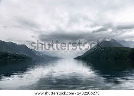 Dramatic clouds cast a majestic reflection on the still waters of a Norwegian fjord, with towering mountains enveloped in mist creating a somber and powerful scene