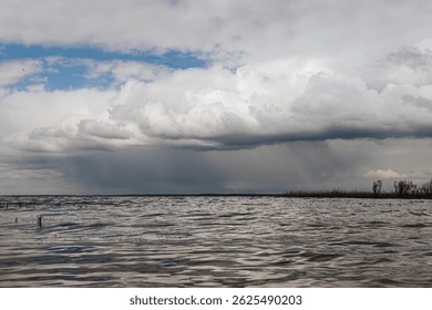 Dramatic clouds loom over a lake shoreline, with distant rainfall visible on the horizon and a hint of blue sky breaking through ample copy space.