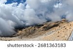 Dramatic clouds rolling over rugged mountain peaks with rocky slopes in sunlight, creating a powerful contrast of earth and sky in a breathtaking alpine view.