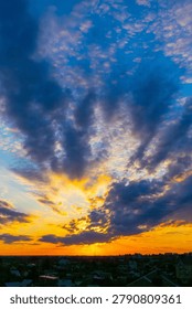 Dramatic clouds stretch wide against golden twilight sky. Sun dips low, casting fiery hues across horizon. Urban skyline silhouetted beneath vast, swirling storm. Colorful sky evokes awe, peace
