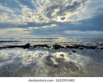 Dramatic clouds and sunlight reflecting in a tidal pool on a peaceful seashore. Evening sky mirrored in wet sand with scattered seaweed.