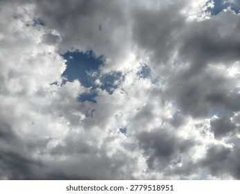 A dramatic cloudscape photograph featuring dense grey and white clouds spreading across the sky with patches of blue visible between them. The textured cloud formations and natural daylight create a dynamic atmospheric scene filled with depth and movement. Captured outdoors, this image is ideal for themes related to weather changes, monsoon season, nature backgrounds, cloudy skies, atmospheric conditions, and scenic sky photography.