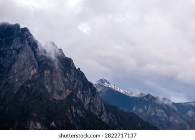 A dramatic, cloudy landscape view of high, rugged mountain peaks in Yunnan, China, with one distant summit partially covered in snow under a heavy, overcast sky.