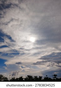 Dramatic cloudy sky with bright sunlight shining through layered clouds above a dark tree line.
