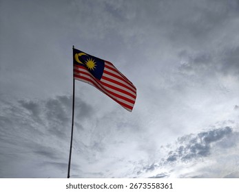 A dramatic cloudy sky frames the Malaysian flag, Jalur Gemilang, flying proudly on a high pole, representing national unity, heritage, and independence day spirit.