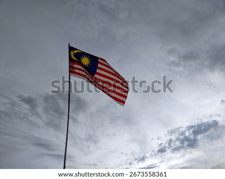 A dramatic cloudy sky frames the Malaysian flag, Jalur Gemilang, flying proudly on a high pole, representing national unity, heritage, and independence day spirit.