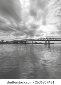 A dramatic cloudy sky looms over a long steel bridge spanning a calm river, with reflections dancing on the water capturing the power of nature and architecture in harmony. Keywords: bridge, river.
