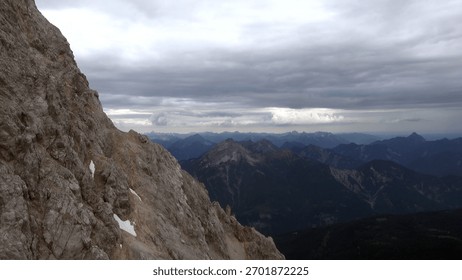 Dramatic cloudy sky looms over rugged mountain peaks, offering a glimpse of a vast natural landscape. Several patches of snow can be seen scattered along the rocky terrain