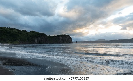 Dramatic cloudy sky over a dark volcanic sand beach with gentle waves and rugged sea cliffs, photographed on the Kamchatka Peninsula, Russia, at dusk