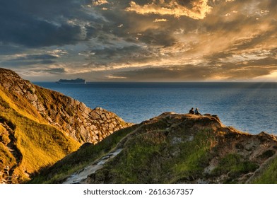 Dramatic cloudy sky over Lulworth Cove with a view of two people watching a peaceful waterscape from the rocky shore, UK