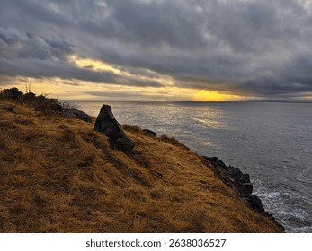 A dramatic cloudy sunrise or sunset over the ocean, viewed from a dry grassy cliffside with a prominent dark rock - Powered by Shutterstock - Get 15% off with code: PIKWIZARD15