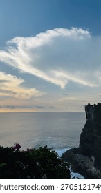Dramatic coastal cliff at sunset with soft golden light, ocean waves, lush greenery, and a silhouette of a small structure against a wide open sky and sweeping clouds.